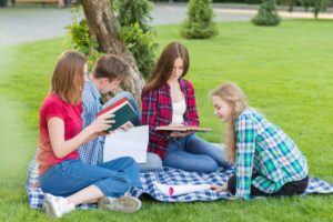 Four students in casual attire gather on a picnic blanket in a park, engaged in reading books and studying outdoors.
