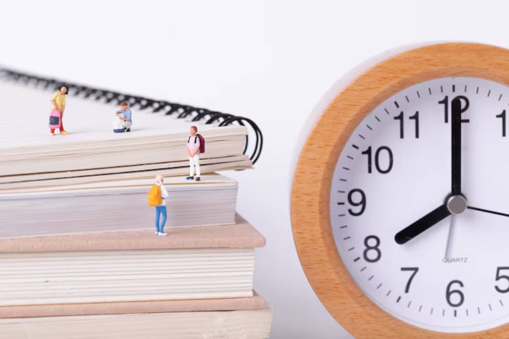 A clock beside stacks of books with miniature figures standing on top, symbolizing time management and academic study.