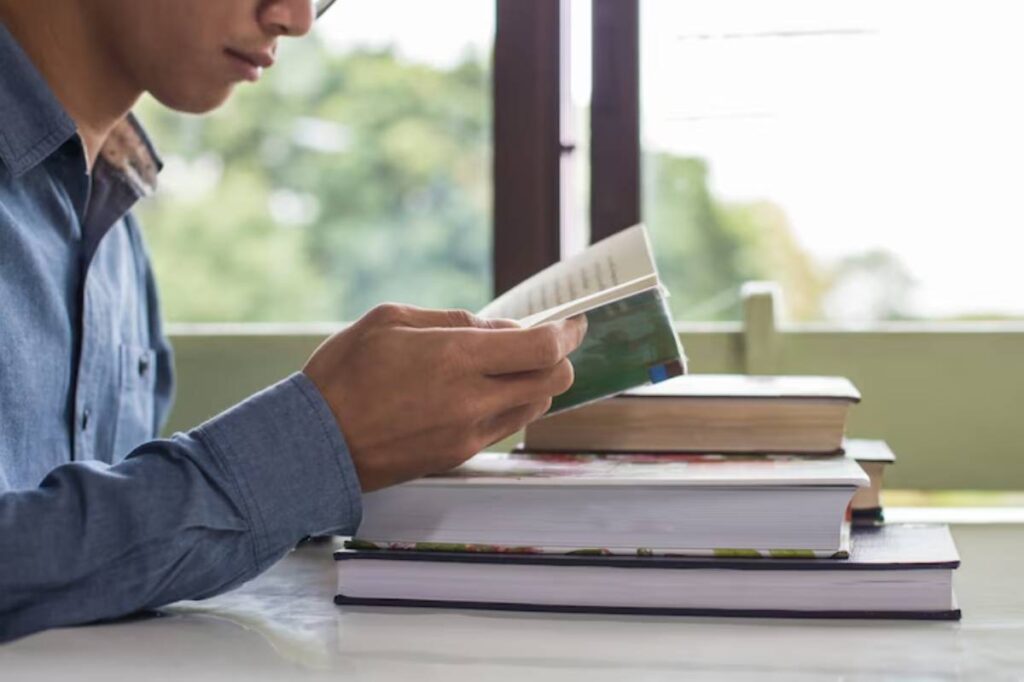 A young man in a blue shirt sits at a table, holding an open book, with several stacked books and a window view in the background.