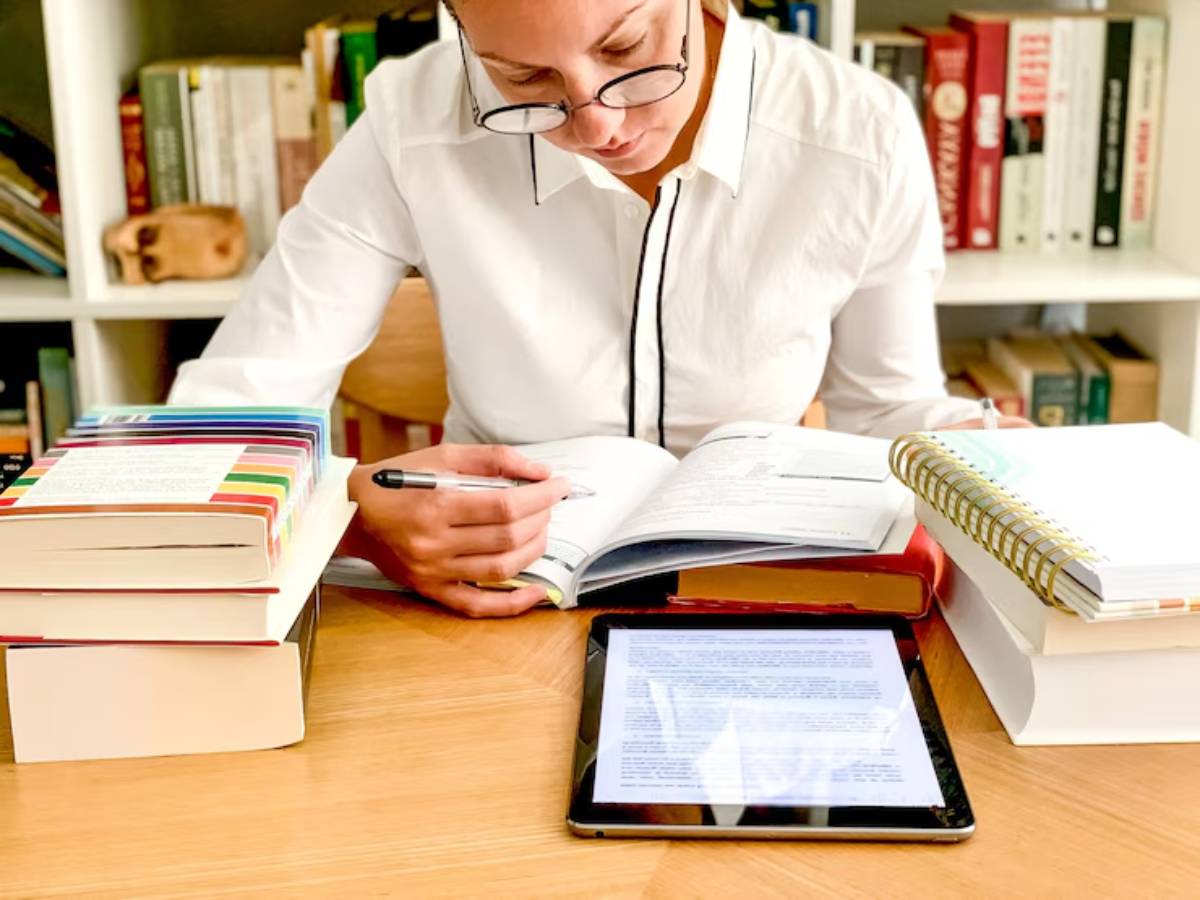 A person studying at a table cluttered with various books, a tablet displaying text, and a notepad, focused on note-taking.