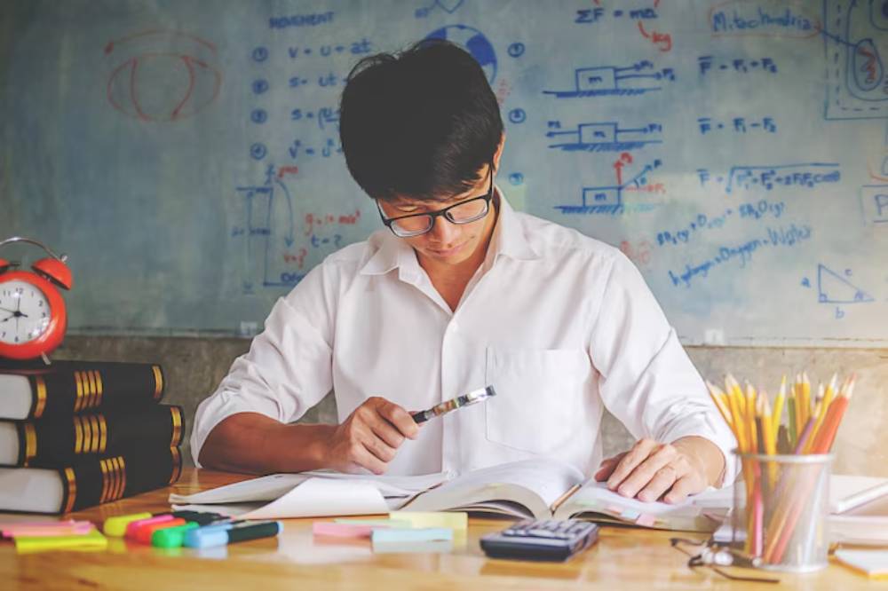 A student in a white shirt studies at a desk filled with books, stationery, and a clock, with educational diagrams on the wall behind.