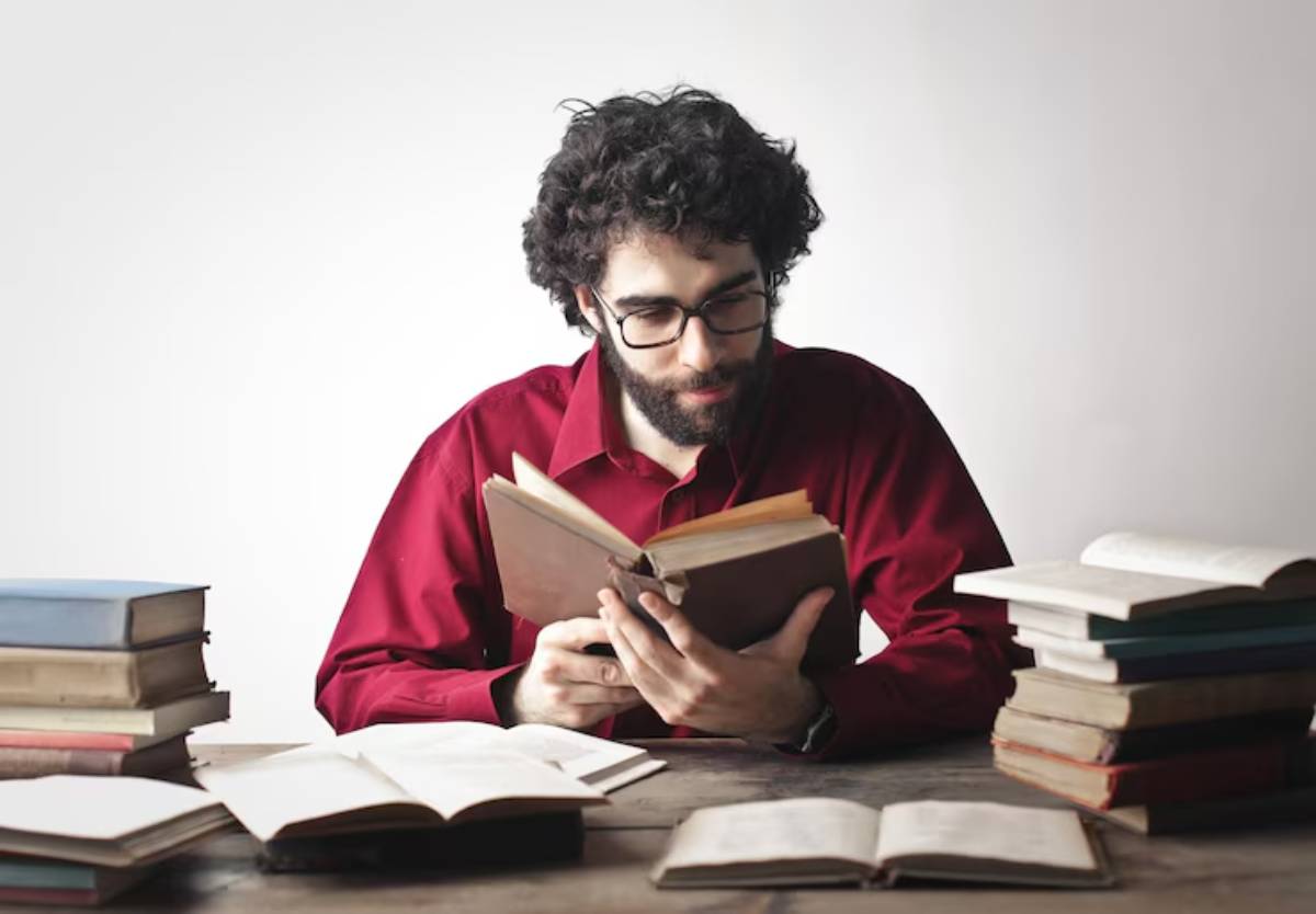 A person wearing a red shirt reads a book, surrounded by stacks of various books on a wooden table, against a light background.