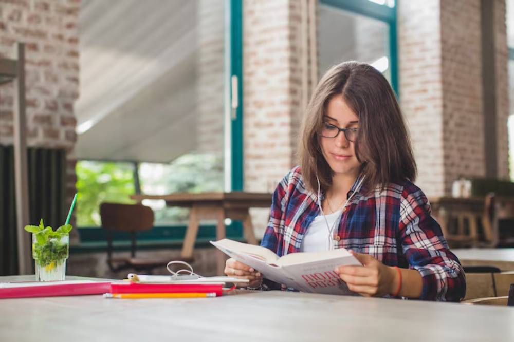 A young woman in a plaid shirt sits at a rustic wooden table, reading a book with a drink and stationery nearby in a cozy café.