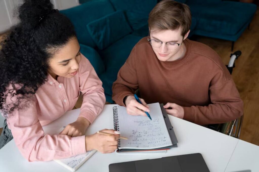 A person in a pink shirt and another in a brown sweater are collaborating at a desk, surrounded by notebooks and a laptop.