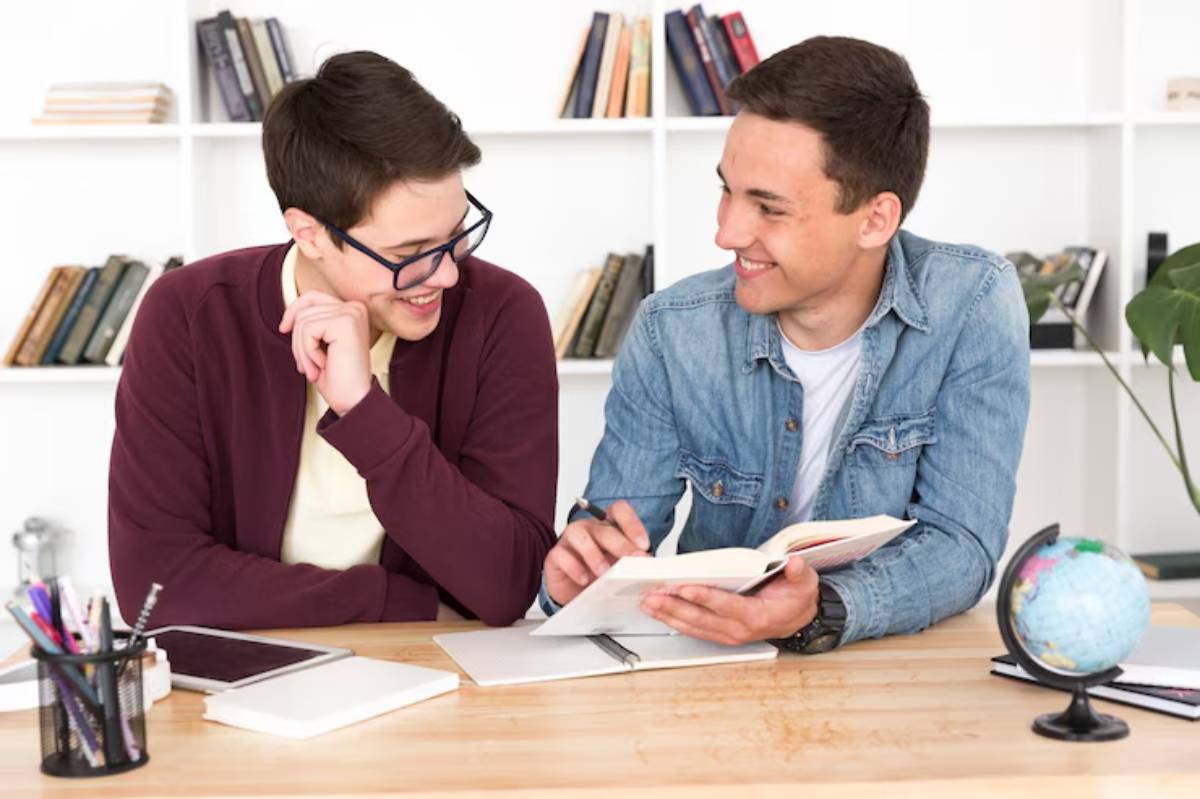 Two young men are studying together at a wooden table, surrounded by books, a globe, and stationery. One reads from a notebook.