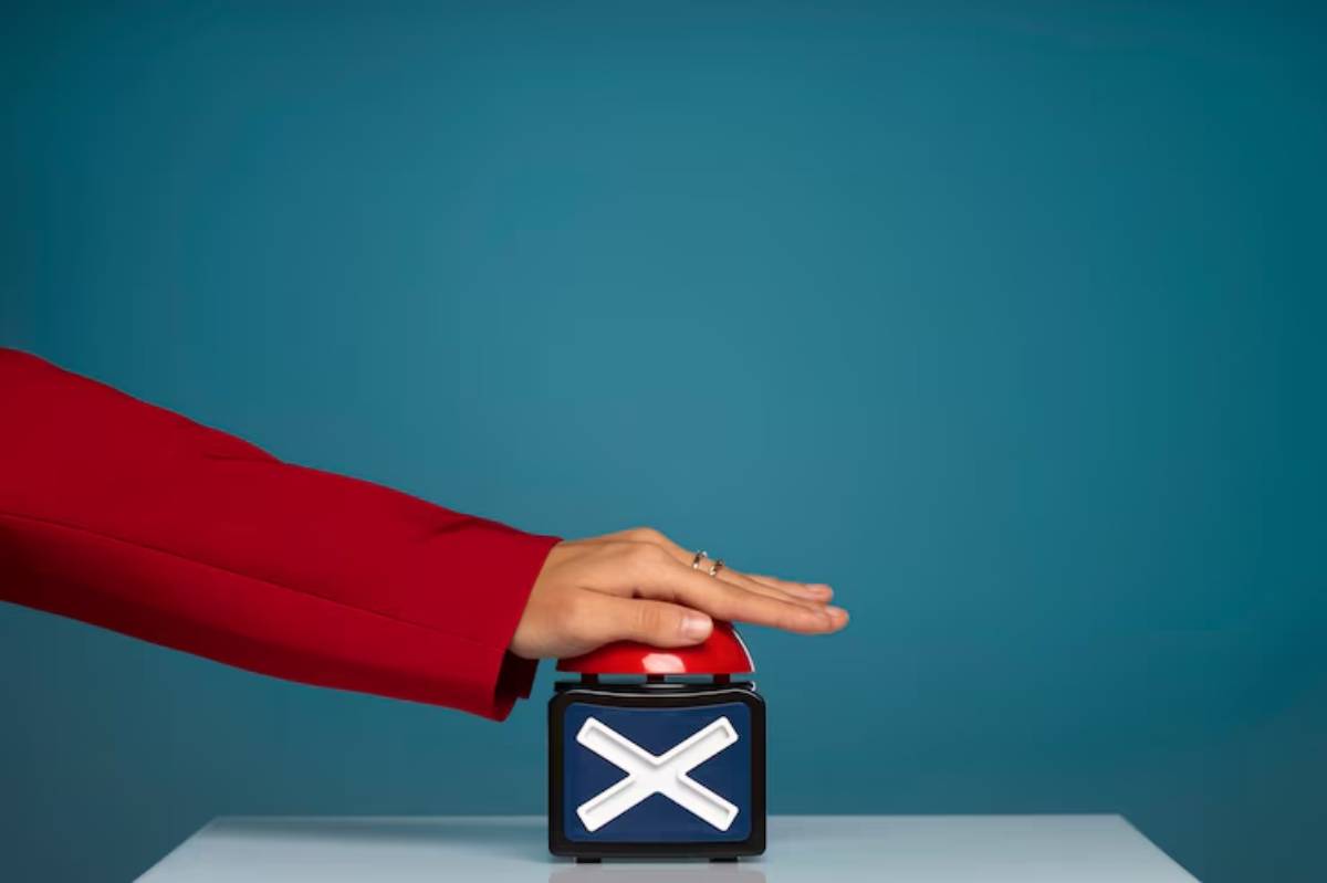 A hand in a red sleeve hovers over a large red button with a white cross symbol, against a solid teal background.