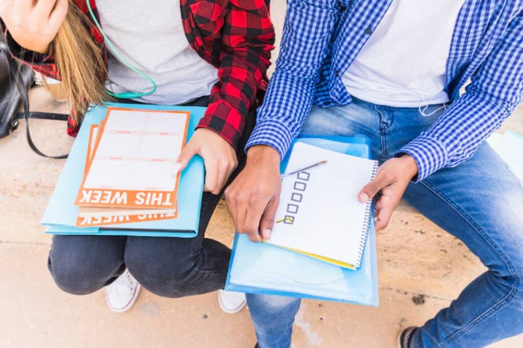 Two students sitting next to each other, studying with notebooks and planners, one wearing headphones, on a stone bench.