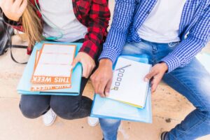 Two students sitting next to each other, studying with notebooks and planners, one wearing headphones, on a stone bench.