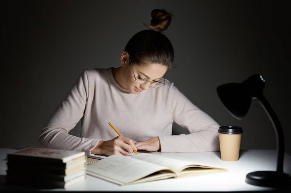 A person sits at a dimly lit table, writing in a notebook, with books piled nearby and a coffee cup beside a desk lamp.