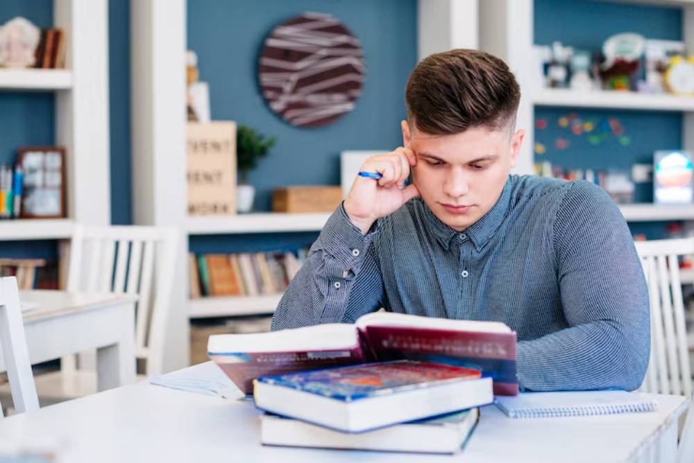 A person sits at a table covered with books and notebooks, deep in thought, in a cozy, well-lit study space with bookshelves.