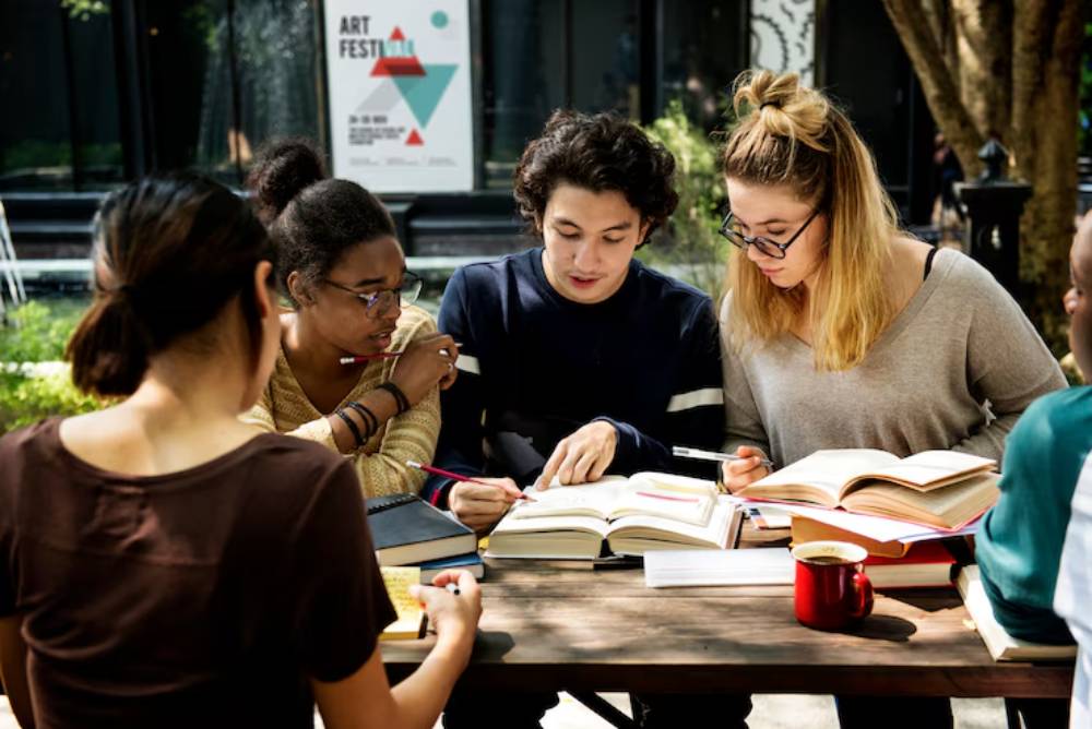 A group of students studies together at a wooden table outdoors, surrounded by books and coffee mugs, under dappled sunlight.