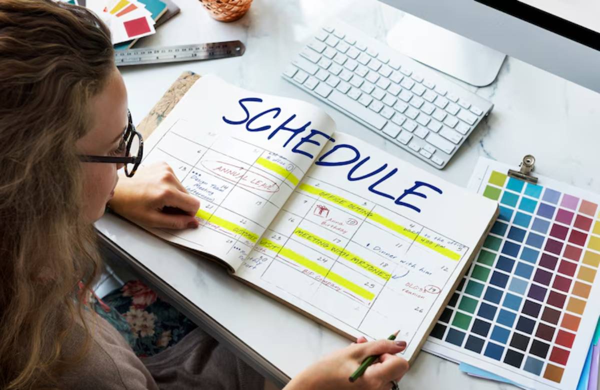 A person writes in a planner titled 'SCHEDULE' with highlighted dates and notes, surrounded by design materials and a keyboard.