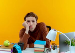 A person sits at a desk cluttered with books, an apple, a coffee cup, and stationery, against a bright yellow wall.