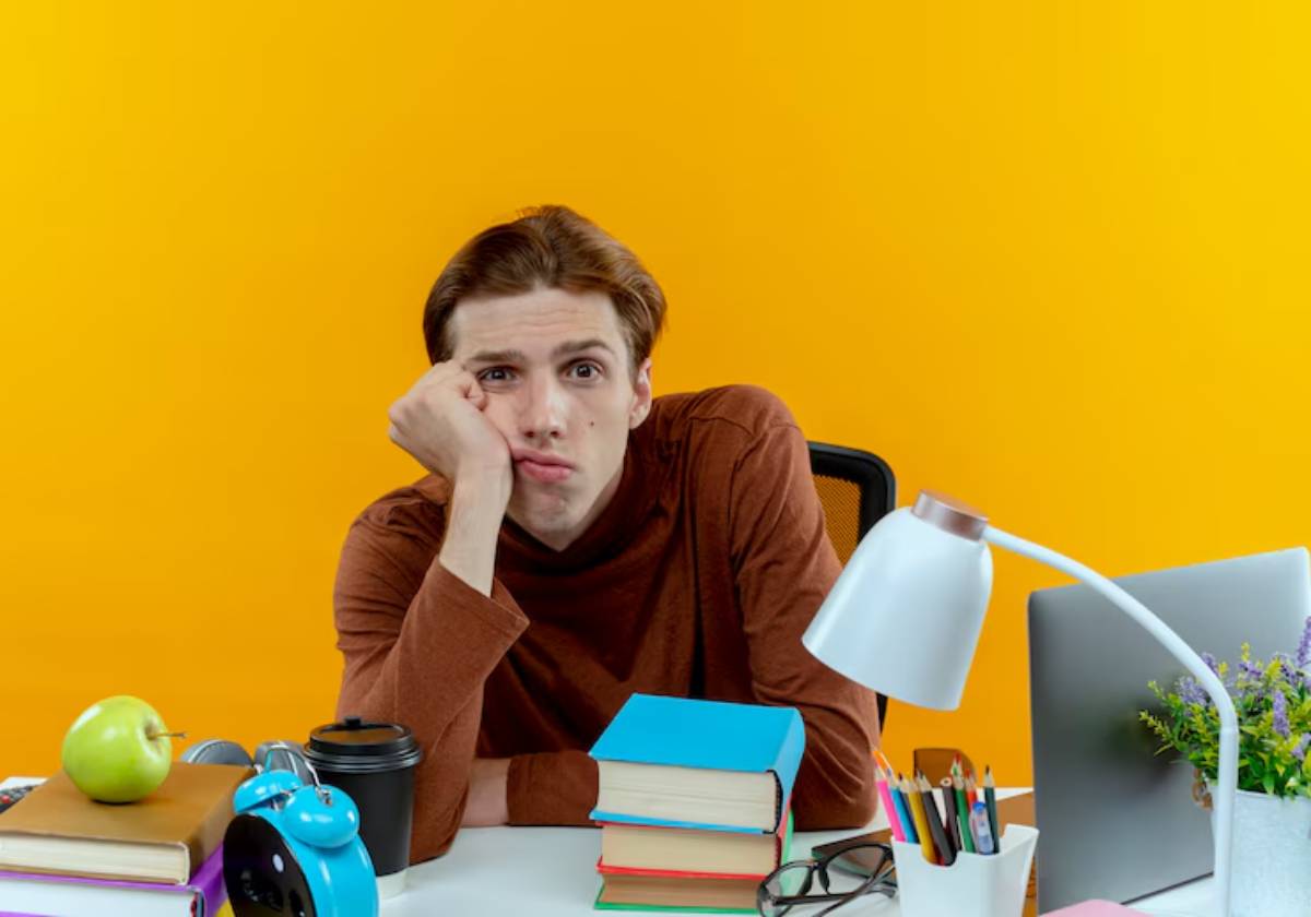 A person sits at a desk cluttered with books, an apple, a coffee cup, and stationery, against a bright yellow wall.