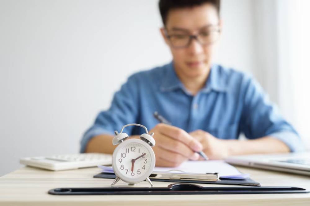 A vintage alarm clock sits on a desk with a person writing notes and a keyboard in the background, emphasizing a productive atmosphere.