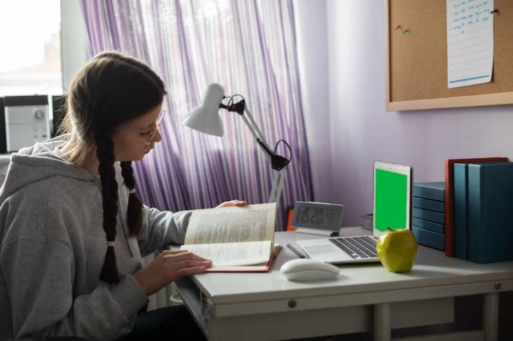 A student studies at a desk, reading a book with a green-screen laptop, clock, lamp, and an apple beside neatly stacked books.