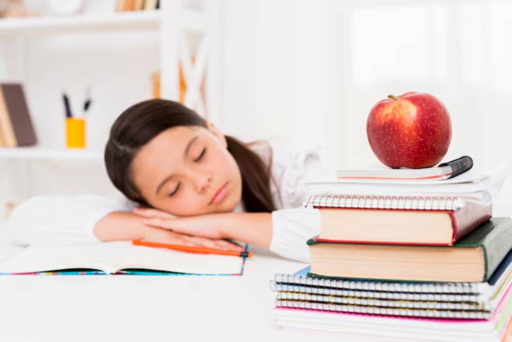 A student rests their head on a desk surrounded by books and a bright red apple.