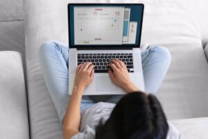 A person types on a laptop, displaying a calendar for November and December, while seated on a light-colored sofa.