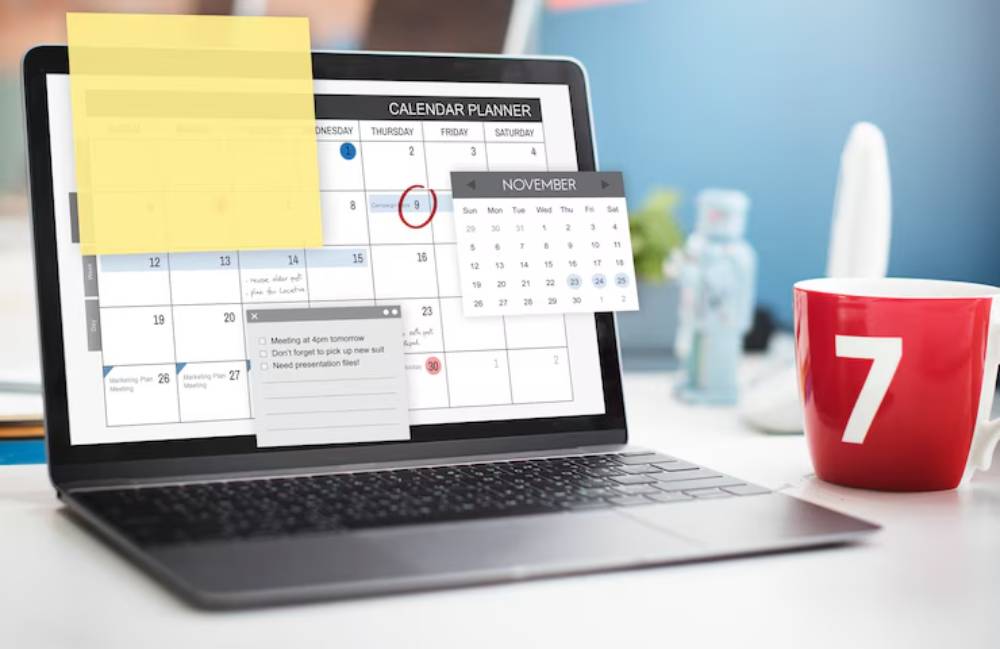 A laptop displays a calendar planner with sticky notes and reminders, next to a red mug with the number 7.