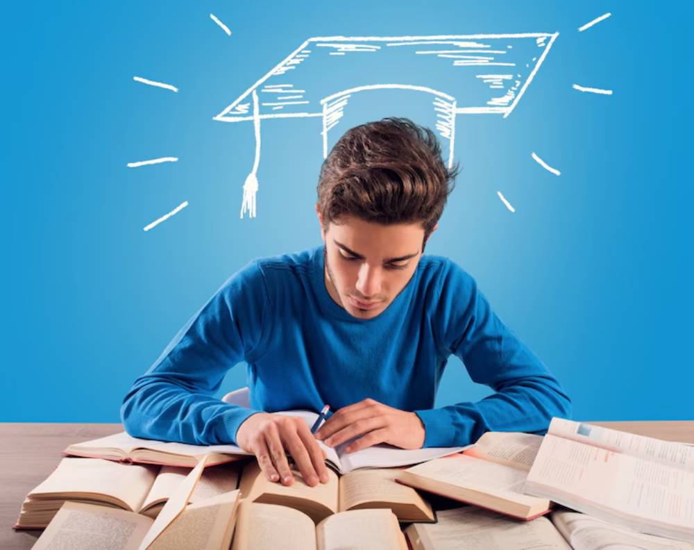 A student in a blue sweater studies intently at a desk filled with open books, with a graduation cap illustration above.