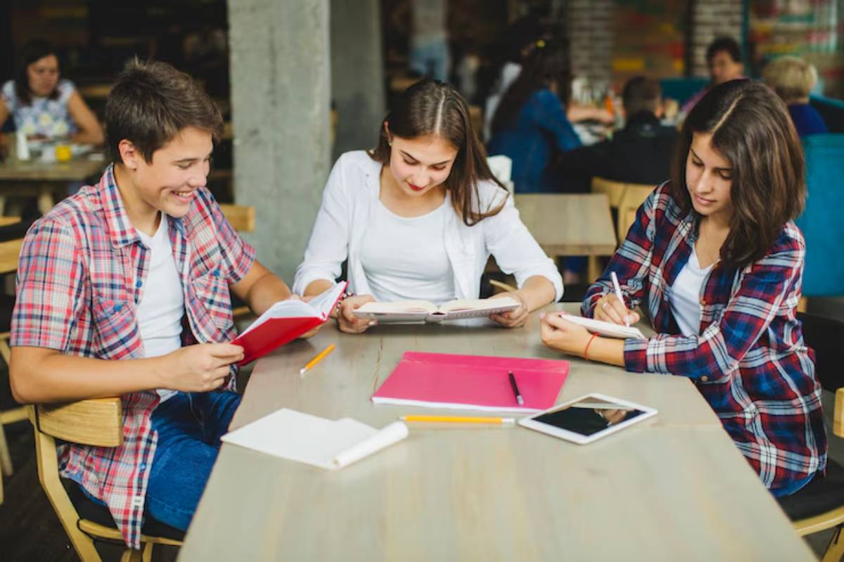 Three students engaged in studying at a wooden table, surrounded by books, notebooks, and stationery in a vibrant café setting.