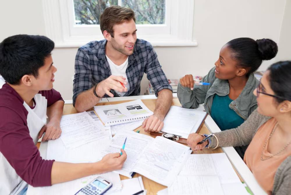 A group of four students engaged in an animated study session around a table filled with notebooks and papers.