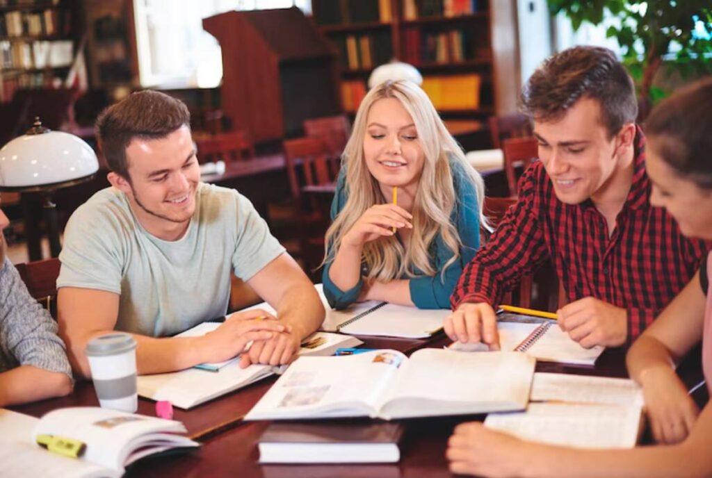A group of students collaborates around a table filled with books and notebooks in a cozy library setting.