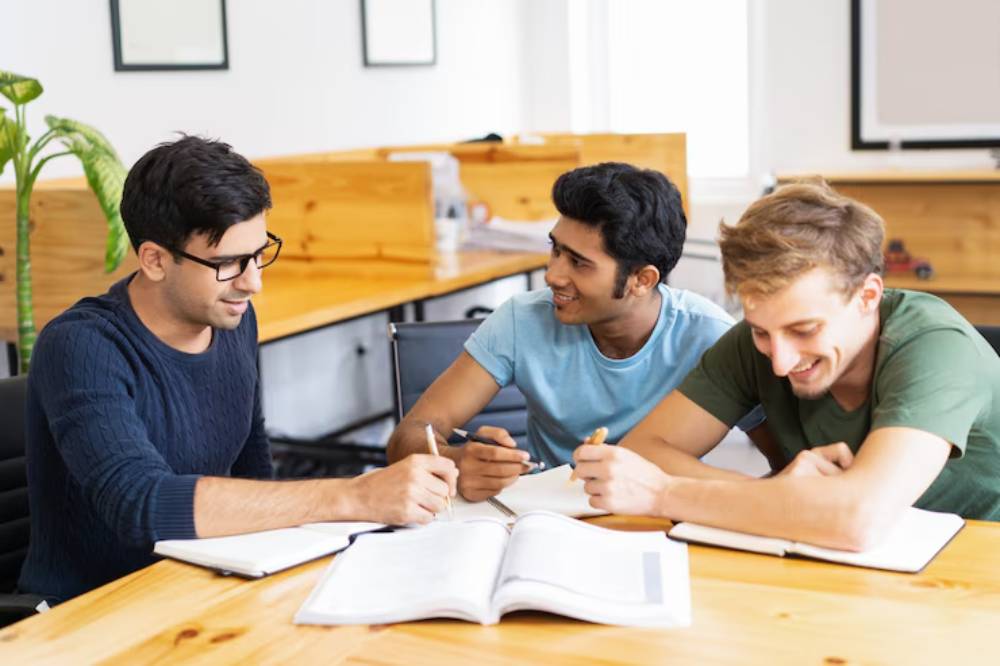 Three individuals are engaged in a focused study session around a table, with notebooks and pens scattered in front of them.