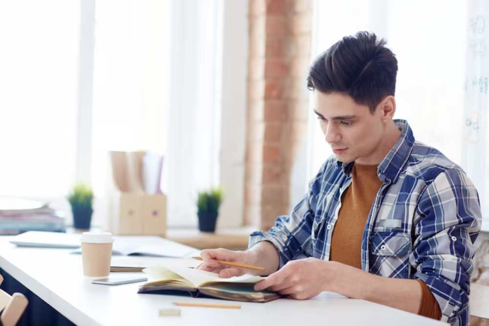 A student in a plaid shirt sits at a desk, studying a book with a coffee cup nearby and sunlight streaming through large windows.