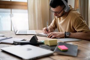 A person wearing headphones writes in a notebook at a wooden desk surrounded by a laptop, notepads, and colorful sticky notes.