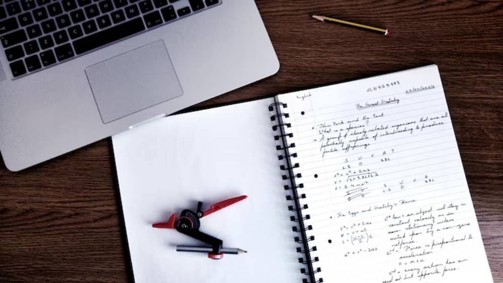 A laptop, notebook with handwritten notes on physics, a compass, and a yellow pencil on a wooden desk.