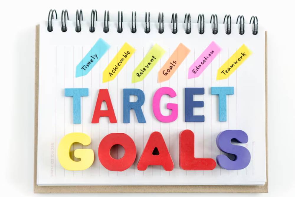 Colorful letters spell 'TARGET GOALS' on a spiral notebook, alongside sticky notes labeled with key goal-setting concepts.