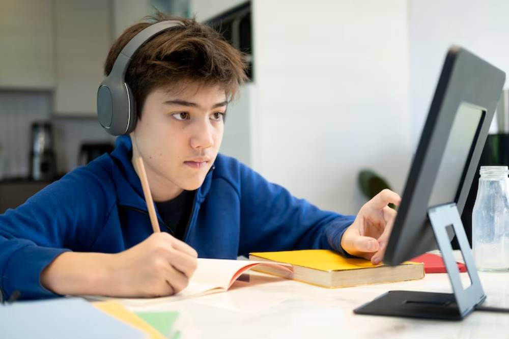 A student in a blue hoodie, wearing headphones, writes notes while following a lesson on a tablet, surrounded by books and a bottle.