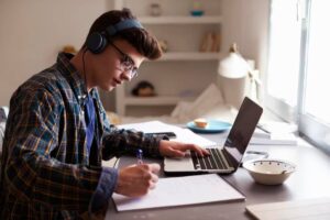 A person wearing headphones learning online on a laptop while writing notes, surrounded by a bowl and a plate of food on a wooden desk.