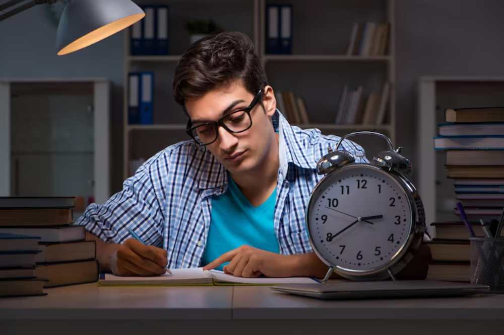 A person studies at a desk surrounded by books, with a clock showing the time in a dimly lit room. A desk lamp provides soft light.