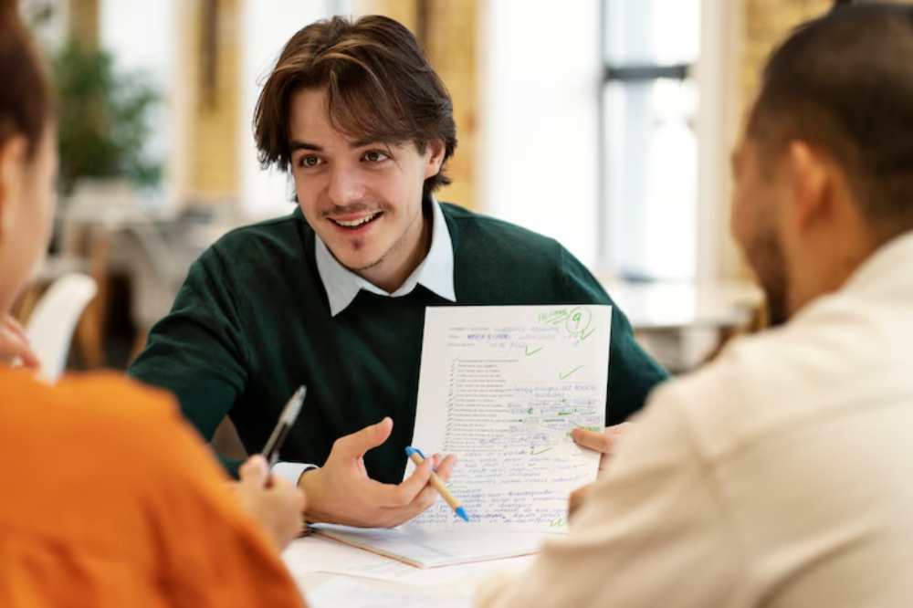 A group discussion at a desk, focusing on a document with notes and checkmarks, as one person gestures while explaining.