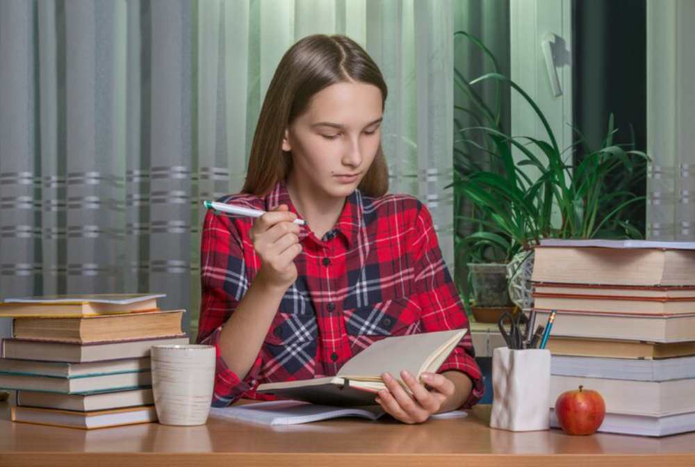 A person in a red plaid shirt holds an open notebook, surrounded by stacks of books, a cup, and an apple on a wooden table.
