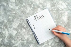A hand holding a pen writes 'Plan' at the top of a blank grid notebook page, with numbered lines for tasks below.