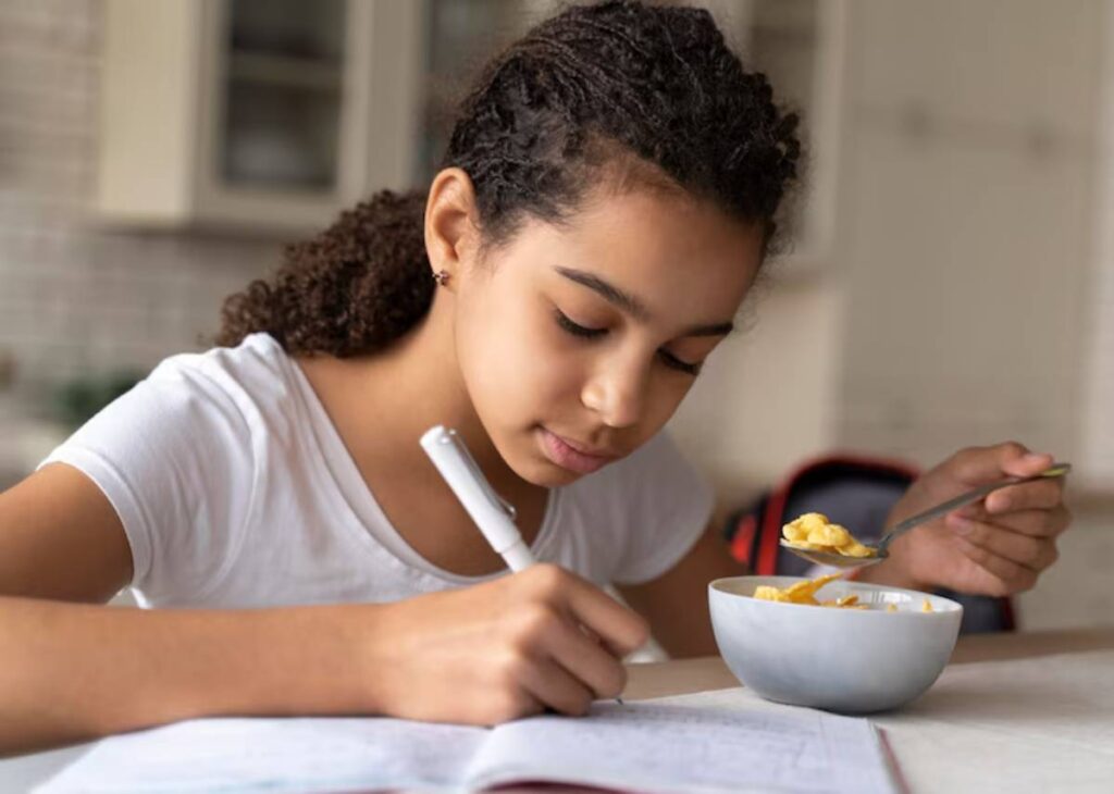A girl sits at a table, enjoying a bowl of cereal while studying, with notebooks and a pen nearby.