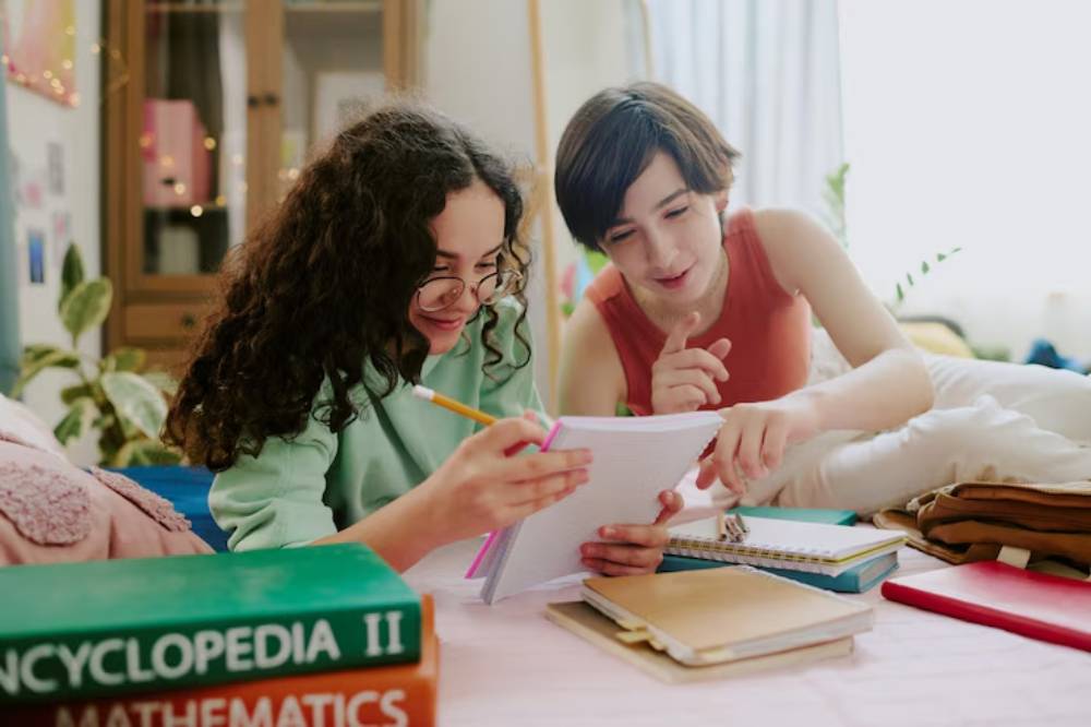 Two individuals are engaged in studying together, surrounded by books and notebooks, in a cozy, well-lit room with plants.