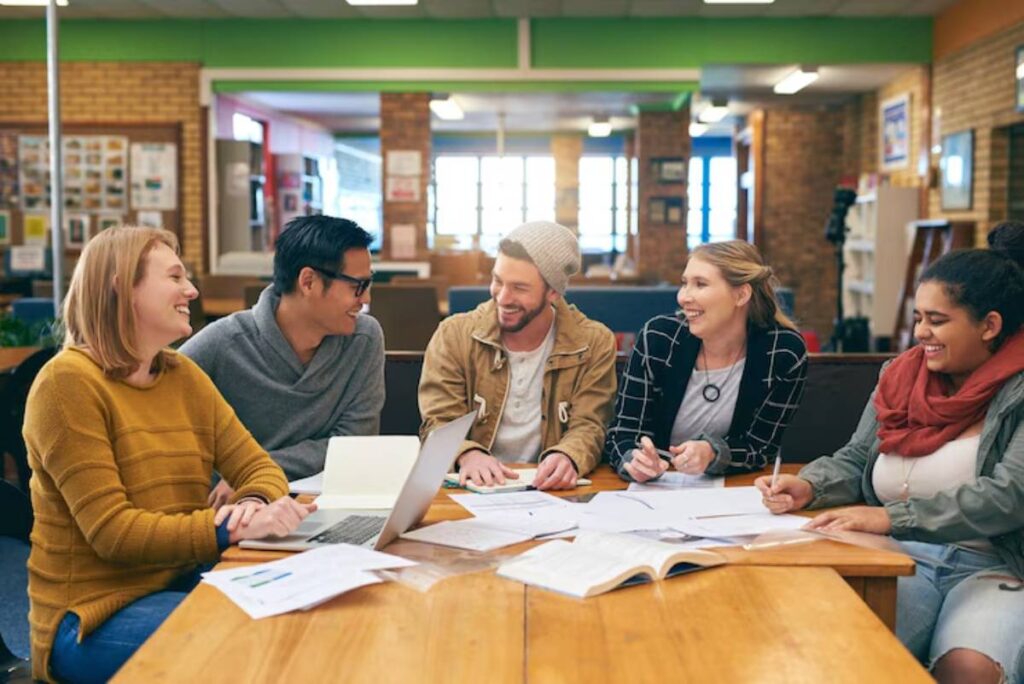 A group of five people collaborate around a table filled with papers and a laptop in a cozy library setting.