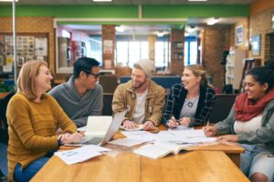 A group of five people collaborate around a table filled with papers and a laptop in a cozy library setting.