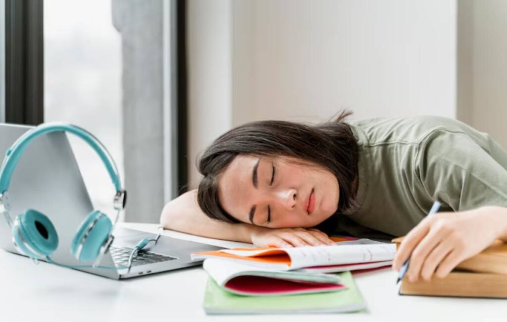 A student rests their head on a cluttered desk, surrounded by books, papers, and a laptop.