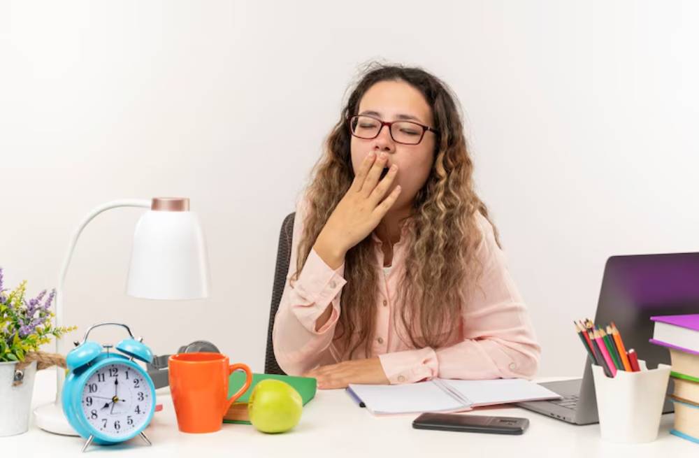 A woman sits at a desk with a lamp, colorful mugs, an apple, an alarm clock, and a laptop, surrounded by books and stationery.