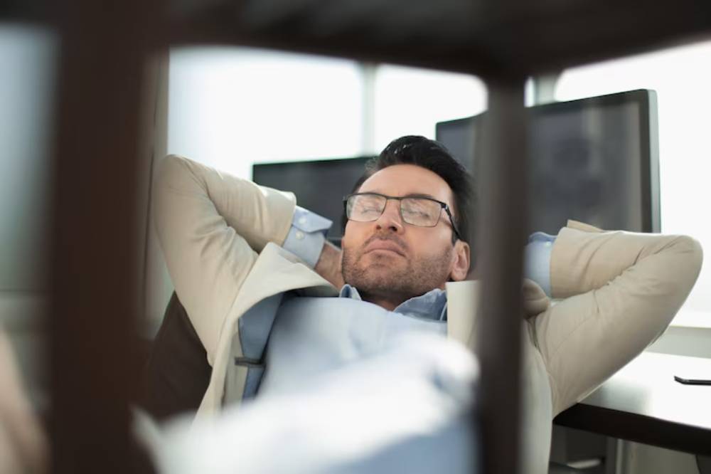 A person in taking nap on a chair, with hands clasped behind their head, in front of computer screens.