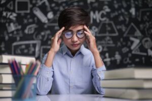 A student in a blue shirt sits at a desk, looking stressed, surrounded by books and colorful pencils against a math-themed blackboard.