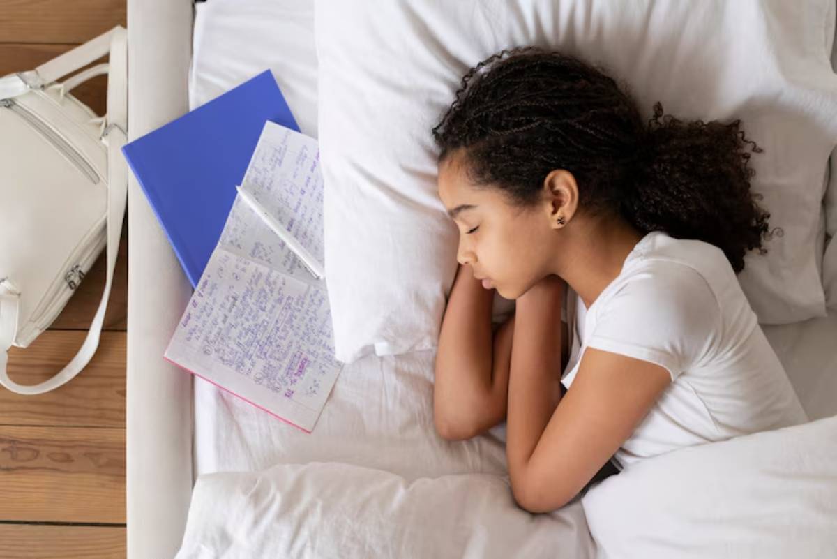 A young person curled up in bed, surrounded by notebooks and a white backpack, suggesting a moment of rest amid study materials.
