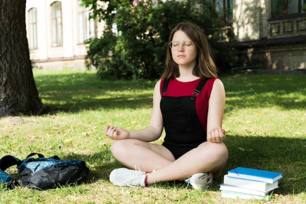 A person sits cross-legged on the grass, practicing meditation, with a backpack and stack of books nearby, under a tree.
