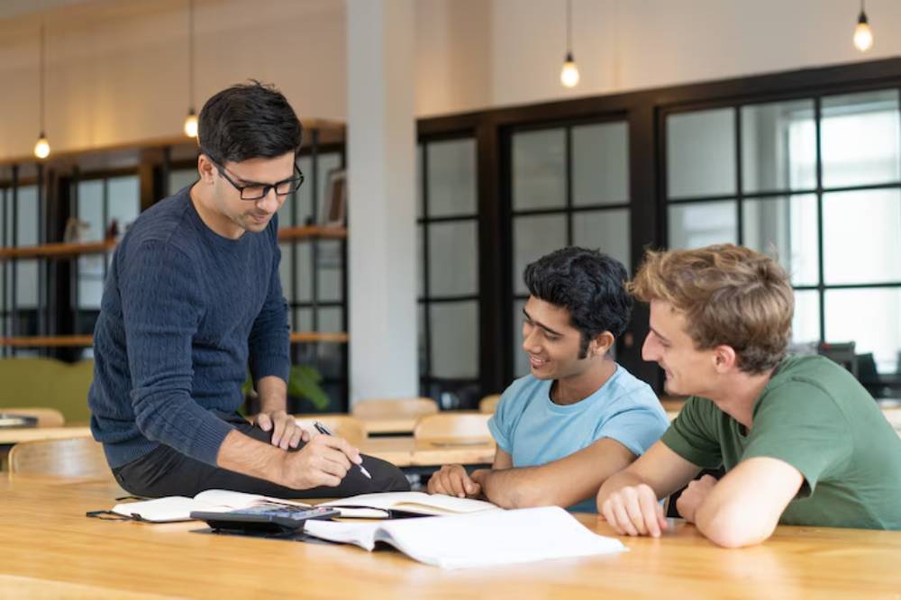 A group of three individuals collaborates around a table covered with notebooks and stationery in a brightly lit study space.
