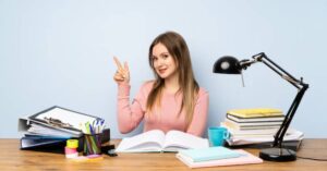 A person in a pink sweater sits at a desk covered in books and stationery, pointing thoughtfully while studying.